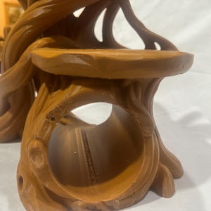 Puede incluir: Un objeto marr&oacute;n hueco con forma de toc&oacute;n de &aacute;rbol con un acabado mate suave. La estructura presenta una amplia abertura circular en la parte delantera y una plataforma plana elevada en la parte superior, con detalles texturizados de ra&iacute;ces.