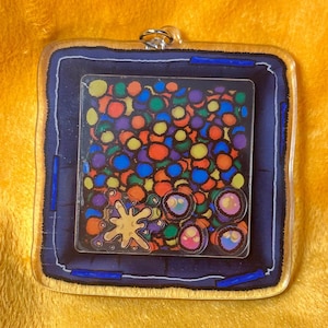 Puede incluir: Un llavero cuadrado de acr&iacute;lico transparente con borde azul y ribete dorado. El interior presenta un dise&ntilde;o colorido de c&iacute;rculos y una salpicadura amarilla.