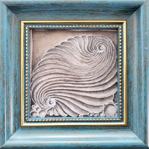 May include: A framed artwork featuring a white, textured, abstract design resembling a seashell. The design is set against a light brown background and framed in a blue and gold frame.