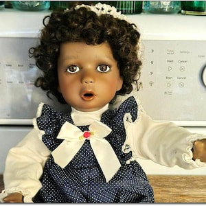 Puede incluir: Una muñeca de piel oscura con cabello castaño rizado que lleva una blusa blanca con ribete de encaje y un vestido de mameluco azul y blanco con lunares con un lazo blanco y un acento de flor roja.