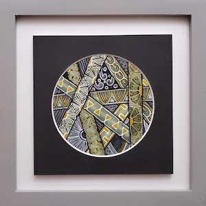 May include: A framed circular artwork with a black background and a white circle in the center. The circle is filled with a geometric pattern of black, white, green, and gold lines and shapes.
