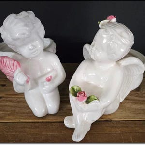 May include: Two white ceramic cherub planter figurines with pink flowers. The cherubs have wings and are kneeling. One cherub is holding a pink flower and the other is holding a pink flower in a green leaf.