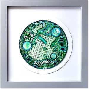 May include: A framed circular watercolor painting with a green and blue abstract design. The painting is surrounded by a white mat and a gray frame. The artist's signature "Laura" is visible in the bottom right corner.