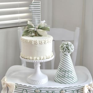 May include: A white frosted cake with a decorative floral border sits on a white pedestal. A striped number one cake topper and a green bow adorn the cake. A matching striped party hat with "Happy Birthday" text and a banner that says "ONE" complete the first birthday theme.
