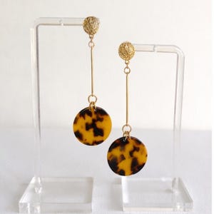 May include: A pair of dangle earrings with a gold-colored post and a textured ball at the top. A thin gold bar connects the ball to a round, tortoiseshell-patterned pendant. The earrings are displayed on a clear acrylic stand.