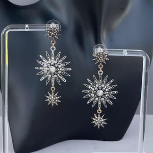 May include: Dangling starburst earrings with a silver and gold finish. Each earring features a large starburst design with clear crystals and a smaller starburst pendant. The earrings are displayed on a black mannequin.