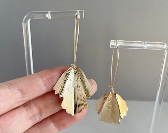 May include: A pair of gold-toned drop earrings featuring a fan-shaped, textured metal leaf design with vertical etched lines. The earrings are suspended from long, thin, curved gold-toned ear wires that extend upward from the top of each leaf.