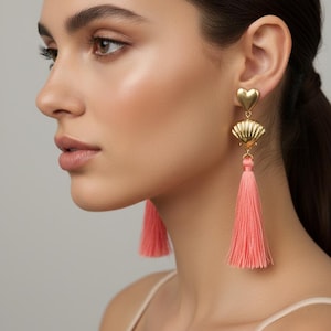 May include: A pair of gold-toned earrings featuring a heart-shaped stud connected to a metallic seashell charm, which suspends a long, vibrant coral-colored thread tassel. The earrings are designed with a post-back closure and hang vertically.