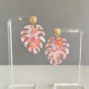 May include: A pair of dangle earrings featuring a leaf design. The earrings have a gold-colored stud and a leaf-shaped pendant with a pink and white marbled pattern. The earrings are displayed on a clear stand against a gray background.