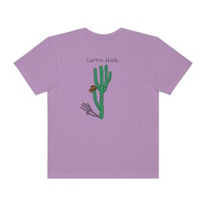 Puede incluir: Camiseta lila con un diseño de cactus verde, un sombrero marrón y el texto "Cactus Hack". El cactus tiene puntos blancos y una sombra. Esta camiseta informal es adecuada para el uso diario.