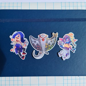 May include: Three holographic stickers on a dark blue notebook. The stickers feature a blue-haired character holding a small object, a gray manta ray with a red and white hat, and a purple character with yellow hair holding a small object.