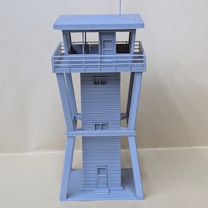 May include: A gray, 3D-printed model of a fire lookout tower featuring a central square structure with textured brick-like walls, a top observation deck with railings, a roof, and a tall antenna mast extending from the top of the roof structure.