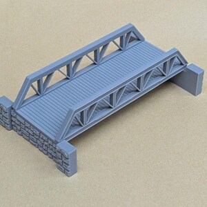 May include: A gray model bridge featuring a planked deck and triangular truss side railings. The structure is supported at each end by rectangular stone-textured abutments, creating a detailed miniature representation of a classic truss bridge.