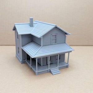 May include: A light gray miniature model of a two-story house featuring horizontal siding, a shingled roof with a chimney, a front porch supported by thin columns, a set of front steps, and multiple rectangular window frames on the exterior walls.