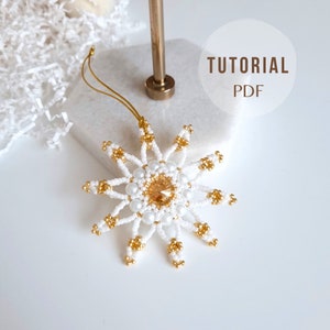 May include: A white and gold beaded snowflake ornament with a clear crystal in the centre. The ornament is hanging from a gold string. The text "TUTORIAL PDF" is visible in the background.