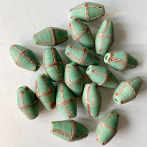 Könnte beinhalten: Eine Sammlung hellgrüner, fassförmiger Perlen mit einem rot-weißen Kreuzmuster. Diese handgefertigten Perlen sind ideal für die Schmuckherstellung und Bastelprojekte. Die Perlen sind etwa 2.5 cm lang.