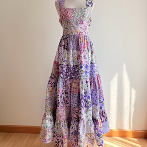 May include: A long, tiered, patchwork dress with a square neckline and thin straps. The dress is made of a variety of floral fabrics in shades of purple, pink, and white.