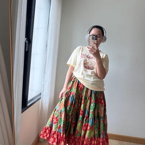Puede incluir: Una camiseta amarilla clara con el texto "TINROSE" y un dise&ntilde;o gr&aacute;fico, combinada con una falda larga escalonada con un vibrante estampado floral de patchwork en rojo, verde y azul. El conjunto se completa con sandalias beige con plataforma.