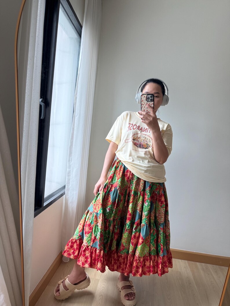 Puede incluir: Una camiseta amarilla clara con el texto "TINROSE" y un dise&ntilde;o gr&aacute;fico, combinada con una falda larga escalonada con un vibrante estampado floral de patchwork en rojo, verde y azul. El conjunto se completa con sandalias beige con plataforma.