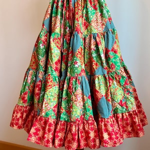 May include: A patchwork skirt with a red drawstring waist. The skirt is made of green, red, and teal fabrics with floral and geometric patterns. The bottom tier has a red ruffle with a gold snowflake design.
