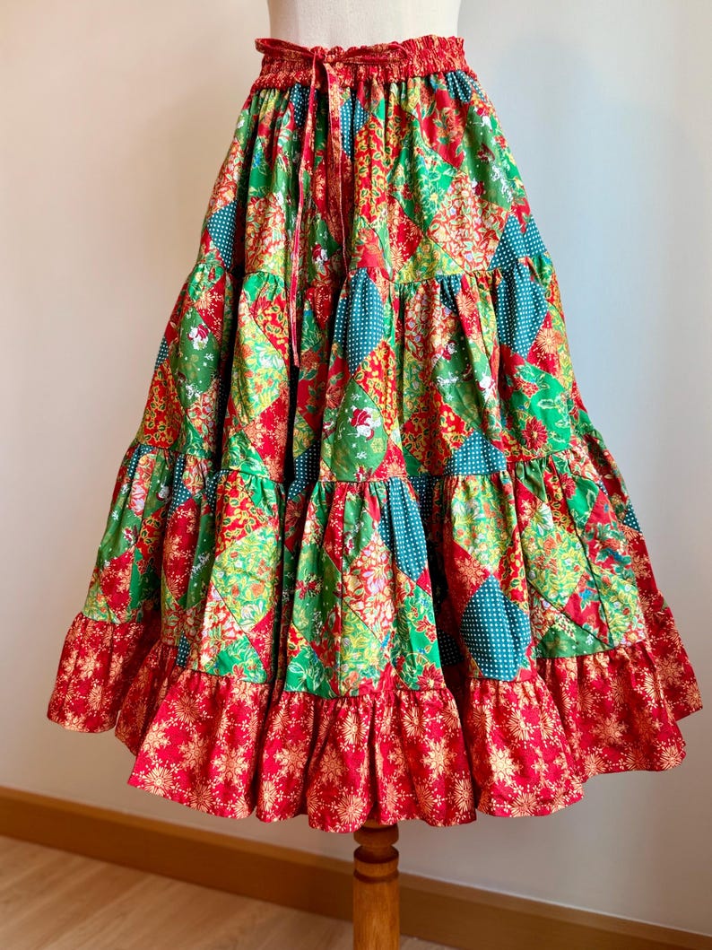 Puede incluir: Falda de patchwork escalonada con cintura roja con cord&oacute;n. La falda presenta una mezcla de telas verdes, rojas y turquesas con estampados florales y geom&eacute;tricos. El dobladillo inferior tiene un volante rojo con un dise&ntilde;o de copo de nieve dorado.
