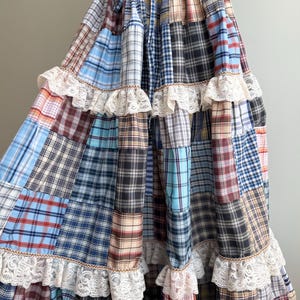Puede incluir: Una falda larga de niveles con un diseño de patchwork de varios patrones de cuadros en tonos azul, marrón, rojo y blanco. La falda tiene una cintura elástica con cordón y está decorada con filas horizontales de encaje color crema.