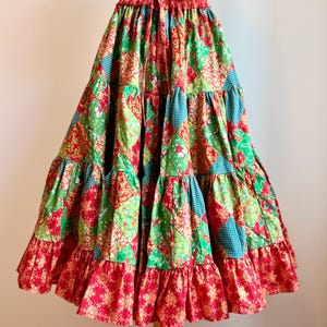 Puede incluir: Falda de patchwork escalonada en tonos rojos, verdes y turquesas. La falda tiene cintura con cord&oacute;n y dobladillo con volantes. La tela tiene estampados florales y geom&eacute;tricos. La falda se exhibe en un maniqu&iacute; de madera. Mide unos 76 cm de largo.