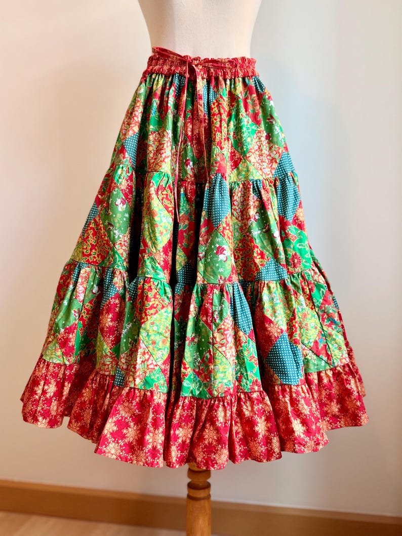 Puede incluir: Falda de patchwork escalonada en tonos rojos, verdes y turquesas. La falda tiene cintura con cord&oacute;n y dobladillo con volantes. La tela tiene estampados florales y geom&eacute;tricos. La falda se exhibe en un maniqu&iacute; de madera. Mide unos 76 cm de largo.