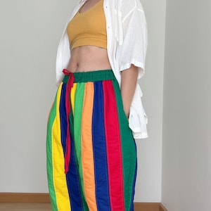 May include: A person wearing a white button-down shirt over a yellow tank top and rainbow striped trousers. They are wearing brown sandals.