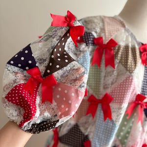 Op de afbeelding: Een witte jurk met een patchwork-ontwerp met verschillende kleuren en stippen. De jurk heeft rode strikken die aan de randen van het patchwork zijn genaaid.