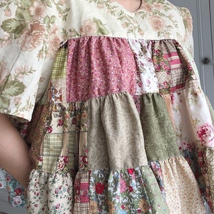 Puede incluir: Una blusa de manga corta de color crema con un diseño de patchwork. La blusa tiene un dobladillo con volantes y está hecha de diferentes telas florales en tonos de rosa, verde y marrón.