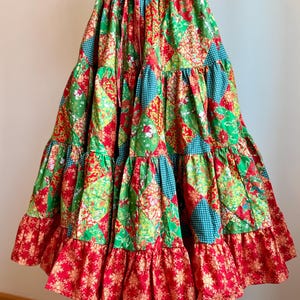 Puede incluir: Falda de patchwork escalonada con cintura roja con cord&oacute;n. La falda presenta una mezcla de telas verdes, rojas y turquesas con estampados florales y geom&eacute;tricos. La parte inferior tiene un volante rojo con un dise&ntilde;o de copo de nieve dorado.