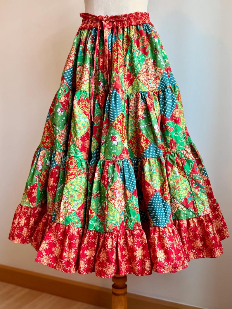 Puede incluir: Falda de patchwork escalonada con cintura roja con cord&oacute;n. La falda presenta una mezcla de telas verdes, rojas y turquesas con estampados florales y geom&eacute;tricos. La parte inferior tiene un volante rojo con un dise&ntilde;o de copo de nieve dorado.