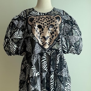 Op de afbeelding: Zwart-wit patroonjurk met een pailletten luipaardgezicht op het lijfje. De jurk heeft korte, pofmouwen en een gelaagde rok. De patronen omvatten zebra strepen en luipaardvlekken.