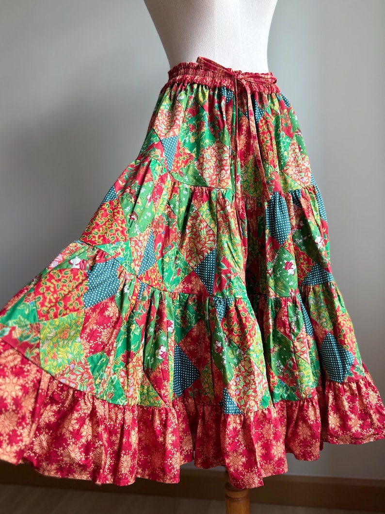 Puede incluir: Falda de patchwork escalonada en tonos rojos, verdes y azules. La falda presenta una cintura con cord&oacute;n y un dobladillo con volantes. La tela tiene estampados florales y geom&eacute;tricos, creando una est&eacute;tica bohemia vibrante.