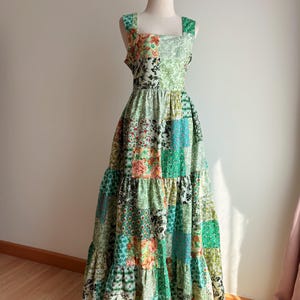 Puede incluir: Un vestido largo de patchwork verde con escote cuadrado y tirantes finos. El vestido tiene una falda de capas con diferentes patrones florales y geométricos.