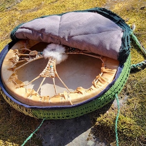 Op de afbeelding: Een ronde, open trommel met een natuurlijke huidkop, met een geweven ontwerp en een pluizig wit accent. De trommel is omhuld in een groen gehaakt frame met een grijze gewatteerde binnenkant en een groen koordhandvat. De trommel rust op een mosachtig oppervlak.