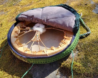 Può includere: Un tamburo rotondo e aperto con una testa in pelle naturale, con un design intrecciato e un accento bianco soffice. Il tamburo è racchiuso in una cornice all'uncinetto verde con un interno imbottito grigio e un manico in corda verde. Il tamburo poggia su una superficie muschiosa.