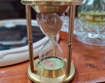 May include: A brass hourglass with a green glass bulb and brown sand. The hourglass is sitting on a wooden table.