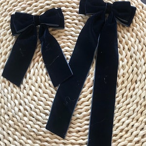May include: Two navy blue velvet hair bows with long, flowing tails. The bows are tied in a classic bow shape, and the velvet material gives them a luxurious look. The bows are placed on a woven, light brown surface.