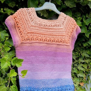 May include: A handmade crochet top with a square neckline and short sleeves. The top features a gradient color scheme, transitioning from peach at the top to purple and then blue at the bottom. The crochet stitch creates an open, airy design.