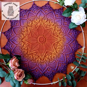 Pode incluir: Uma toalha de mesa de crochê feita à mão em um padrão de estrela, com um gradiente de cores de laranja a roxo. A toalha de mesa é exibida em um aro branco, com o logotipo "Crochet Frog Pattern Designer" no canto superior esquerdo. Rosas e vegetação circundam a toalha de mesa.