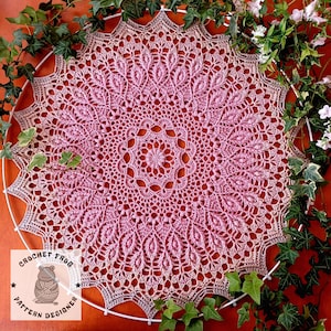 Puede incluir: Un tapete de ganchillo circular hecho a mano en tonos rosa y beige, con un patrón floral detallado. El tapete está colocado sobre un aro blanco y rodeado de hiedra verde y flores blancas. Se ve el logo Crochet Frog Pattern Designer.