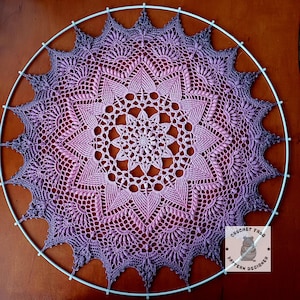Puede incluir: Un atrapasueños circular hecho a mano con un diseño de ganchillo detallado. El patrón de ganchillo presenta un degradado de colores, que va desde un centro rosa claro hasta tonos morados en los bordes. Montado en un aro de metal blanco.
