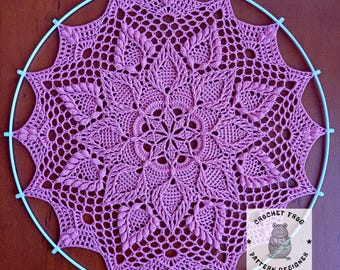 May include: A pink crochet doily with a delicate floral design. The doily is displayed on a blue metal hoop. The text "Crochet Frog Pattern Designer" is visible in the lower right corner of the image.