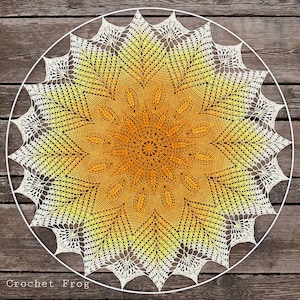 May include: A white crochet doily with a yellow and orange ombre design. The doily is circular and has a delicate lace pattern. The text "Crochet Frog" is visible in the bottom left corner of the image.