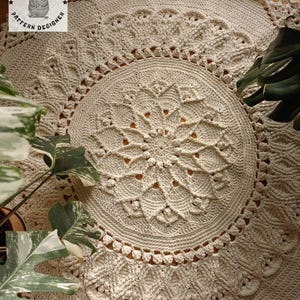 Può includere: Un grande tappeto rotondo all'uncinetto di colore crema con un motivo dettagliato e intricato. Il tappeto ha un design circolare con un motivo centrale e più strati di pizzo all'uncinetto.