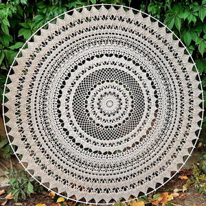 Op de afbeelding: Een groot, rond gehaakt doily met een delicaat, ingewikkeld patroon in beige en witte tinten. Het doily hangt aan een muur met groen gebladerte op de achtergrond.