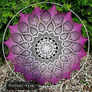 Op de afbeelding: Een groot, rond gehaakt doily met een wit centrum en een verloop van paars naar roze aan de randen. Het doily wordt getoond op een witte metalen ring.