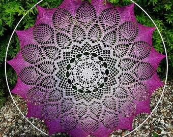May include: A large, circular crochet doily with a white center and a gradient of purple to pink around the edges. The doily is displayed on a white metal hoop.